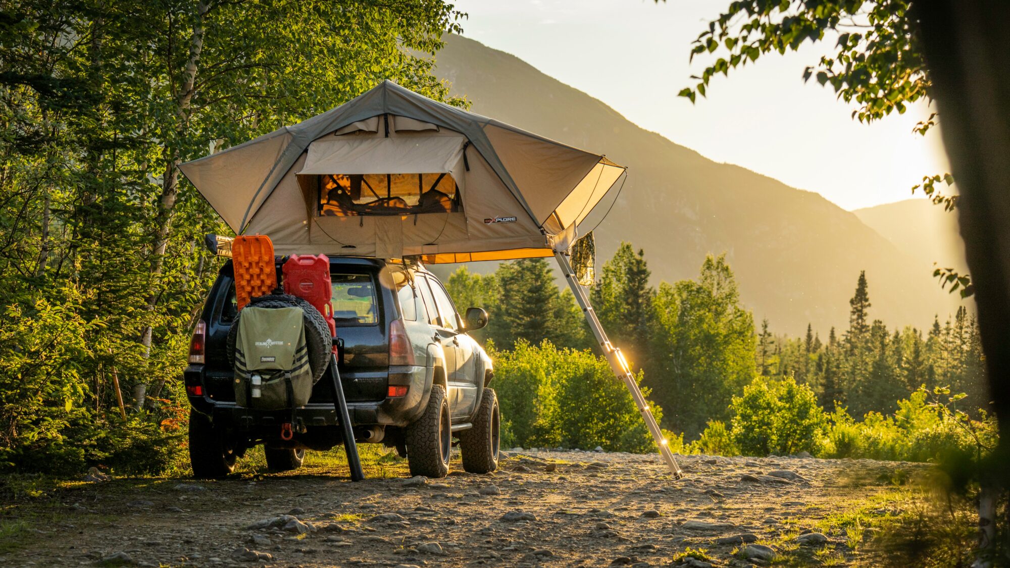 A car is parked in a scenic forest setting with a rooftop tent extended. The sun is setting, casting a warm golden light on the scene. The tent is mounted on the roof of the car and has an awning for extra shade. The car is equipped with outdoor gear including backpacks and storage containers. Surrounding the campsite are lush green trees and mountains in the background, creating a serene and picturesque outdoor environment.