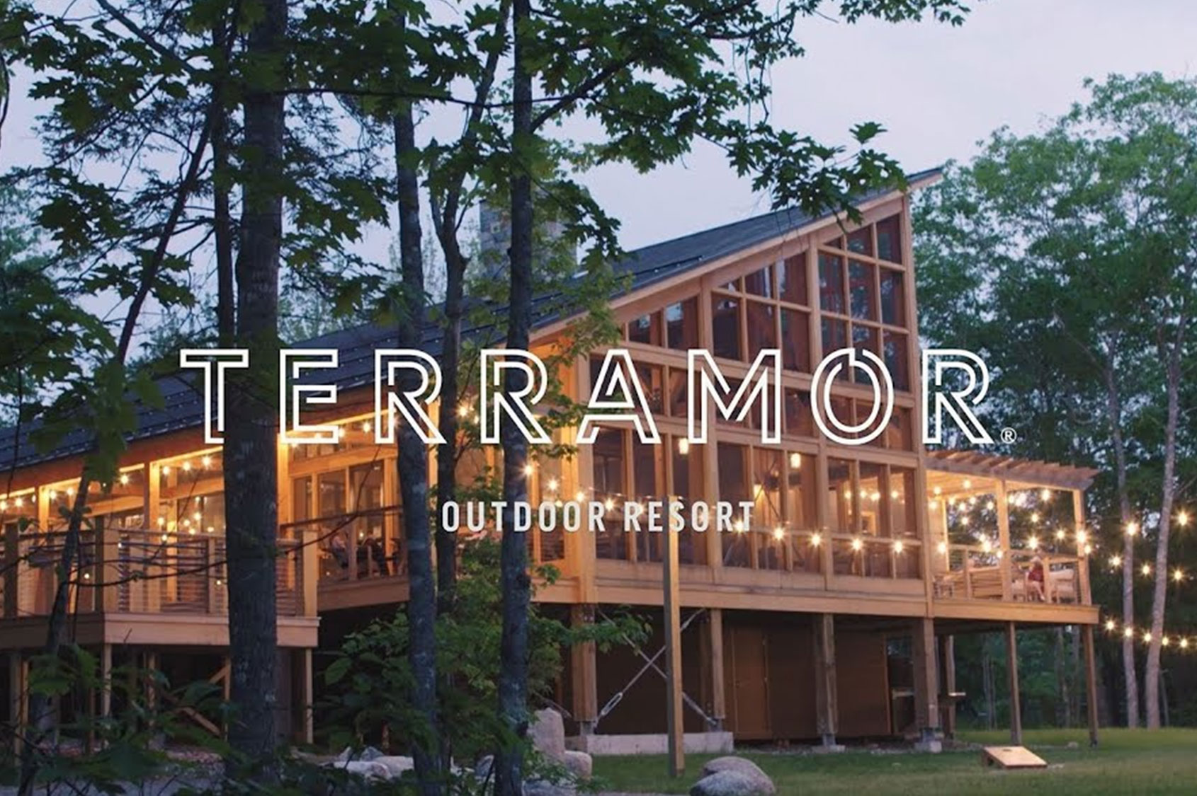 The image shows an evening view of the Terramor Outdoor Resort, featuring a large, modern wooden building with expansive windows and a spacious balcony. The building is lit with warm string lights, creating a cozy and inviting atmosphere. The surrounding area is lush with trees and greenery, adding to the serene and natural setting of the resort.