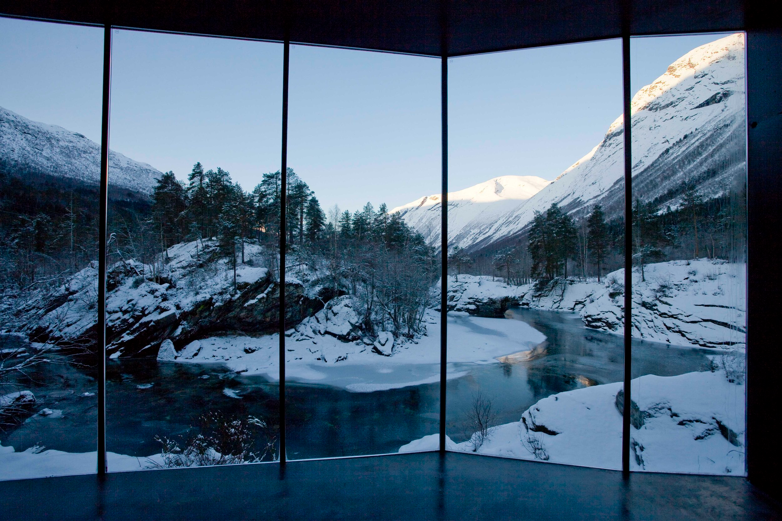 A breathtaking winter view from a glass-walled retreat overlooking a serene, partially frozen river and snow-covered mountains. This immersive landscape hotel experience offers a seamless connection to Norway’s pristine wilderness. Blog post by Sage Outdoor Advisory, offering Glamping and RV Feasibility Studies
