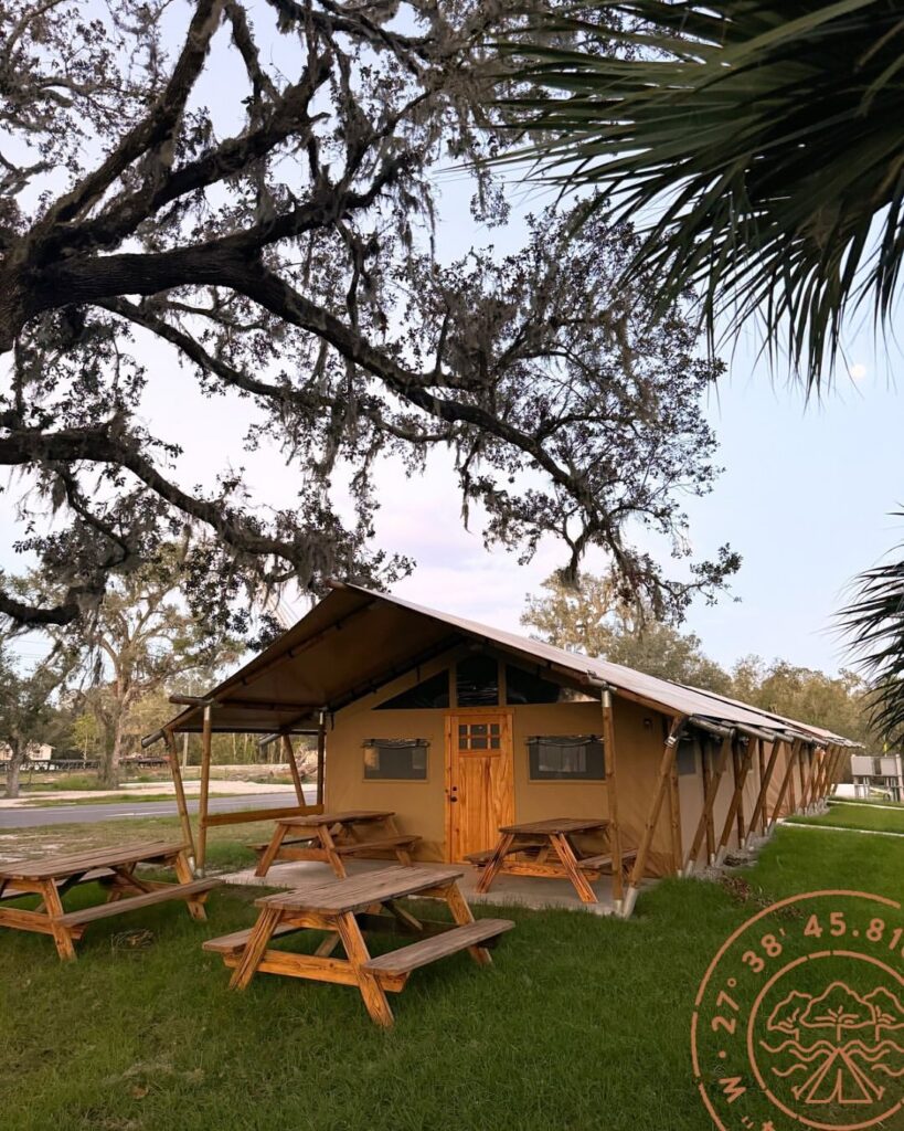 A spacious glamping tent at Peace River Oaks with multiple picnic tables on a grassy lawn, offering a communal outdoor setting. The tent has a wooden door and canvas walls, blending rustic charm with modern amenities. Spanish moss drapes over large oak trees, adding to the scenic atmosphere. Sage Outdoor Advisory provides data-driven insights to support the expansion and success of outdoor hospitality businesses.





