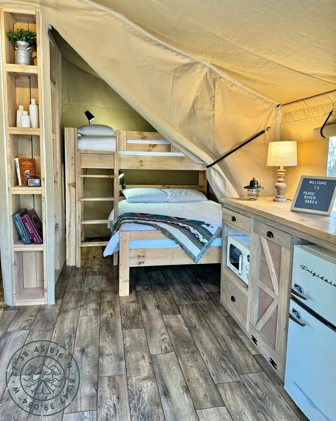 A cozy glamping tent interior at Peace River Oaks, featuring a wooden bunk bed, a neatly made double bed with a decorative blanket, and a kitchenette with rustic cabinetry. A warm lamp sits on the counter, adding to the inviting atmosphere. A wooden bookshelf holds small decor items and books. Sage Outdoor Advisory provides expert insights to help outdoor hospitality businesses like Peace River Oaks thrive.