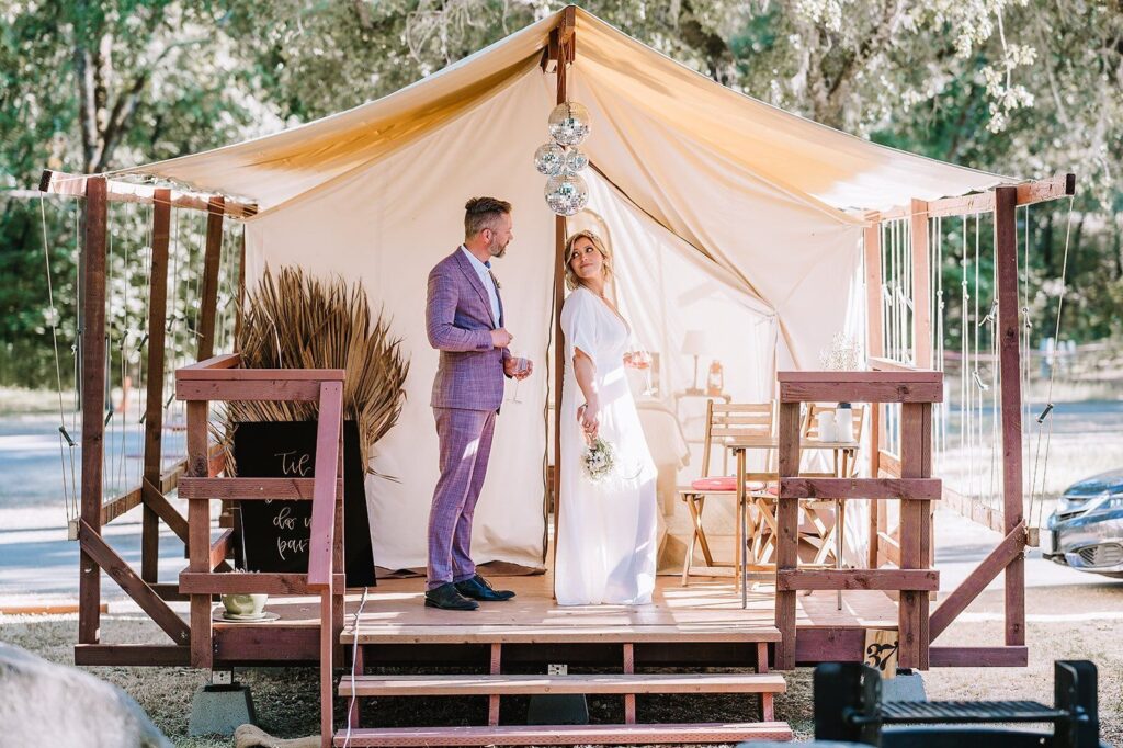 A couple dressed in wedding attire stands outside a stylish glamping tent, decorated with hanging disco balls and rustic furnishings. The groom, in a lavender suit, and the bride, in a flowing white dress, share a moment in the golden outdoor light, blending romance with a luxury outdoor experience. Sage Outdoor Advisory assists glamping operators in optimizing their sites for events and enhancing profitability through strategic planning and data-driven consulting.