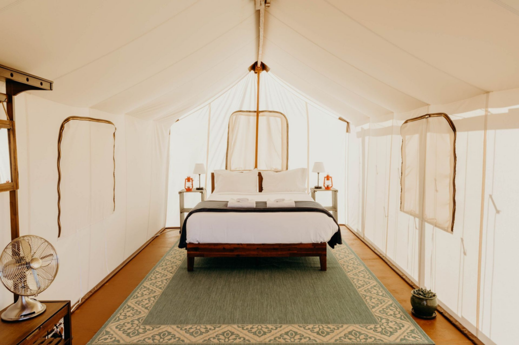 A spacious glamping tent interior with a plush queen-sized bed, soft linens, and rustic nightstands with lantern-style lamps. The light-colored canvas walls and natural wooden flooring create a bright, airy, and inviting atmosphere for a peaceful nature escape. Sage Outdoor Advisory provides expert guidance on market positioning, operational efficiencies, and investment strategies to help outdoor lodging businesses thrive.