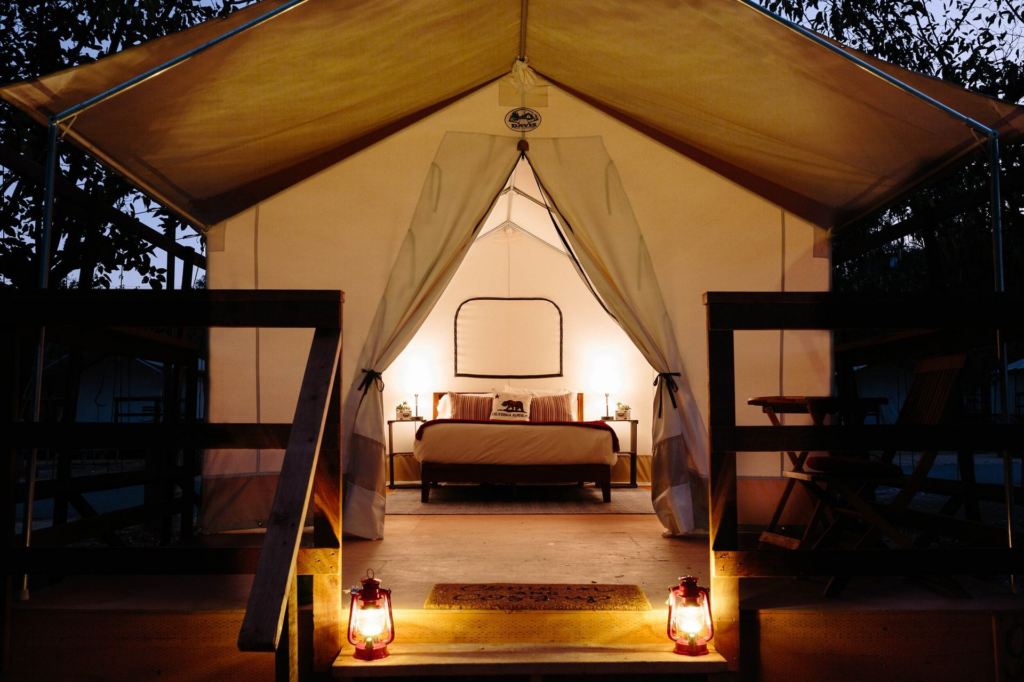 A glamping tent illuminated by warm lighting and vintage lanterns at night. The canvas tent, framed by a wooden deck, features a comfortable queen-sized bed, soft bedding, and cozy ambiance, creating a serene nighttime retreat under the stars. Sage Outdoor Advisory supports outdoor hospitality businesses in designing guest experiences that drive higher occupancy and revenue through feasibility studies and operational insights.