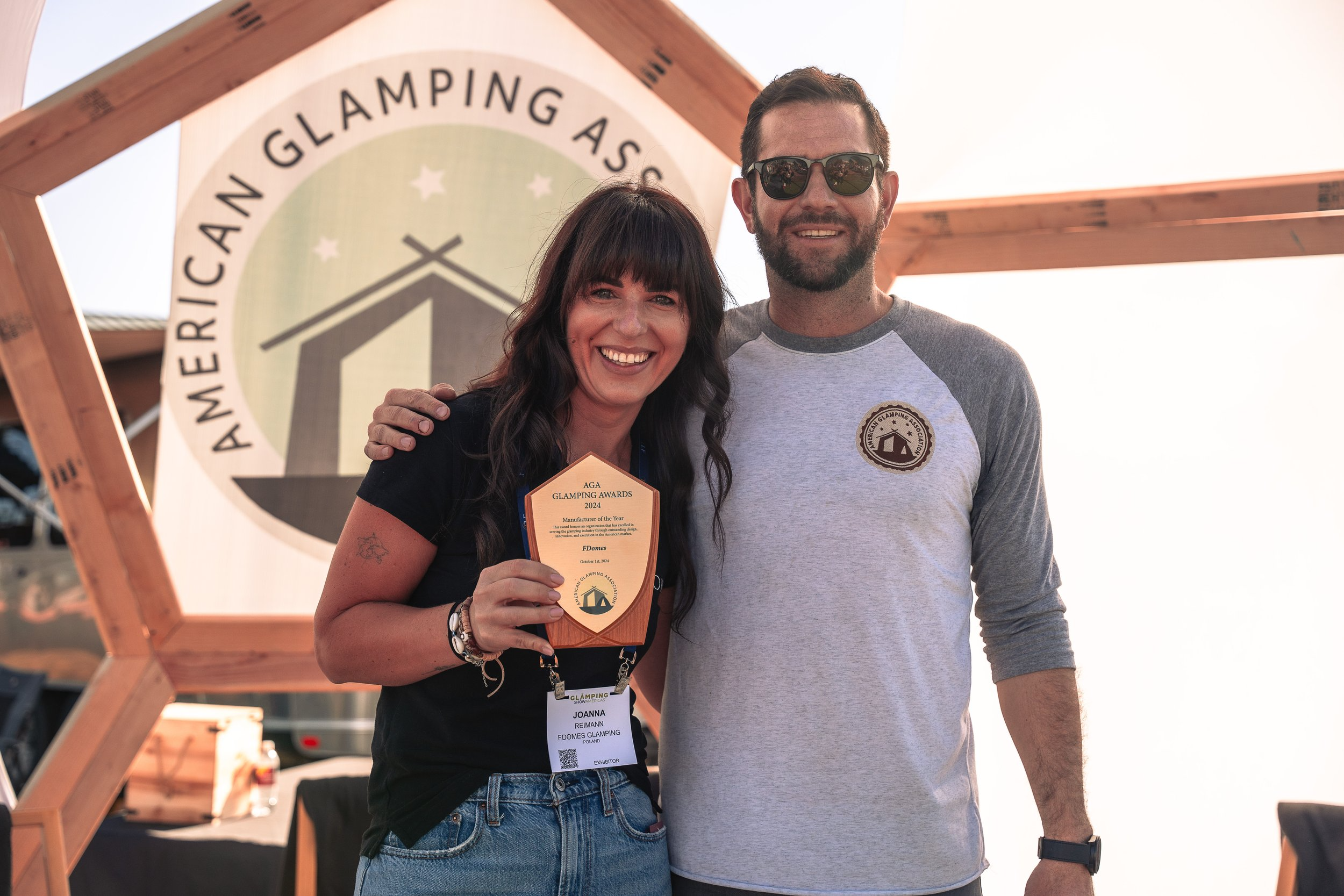 Ruben Martinez, Founder of the American Glamping Association, poses with Joanna Rembialkowska of FDomes Glamping, who is smiling and holding the “Manufacturer of the Year” award at the AGA Glamping Awards 2024. They are standing in front of a large AGA banner at an outdoor event.