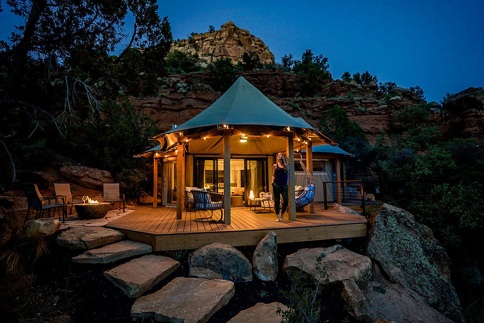 A luxury glamping tent nestled among the red rock cliffs of southern Utah at dusk, illuminated by soft lights and surrounded by natural rock steps and desert vegetation. A guest stands on the wooden deck, enjoying the serene setting. This glamping destination exemplifies high-end outdoor hospitality design. Featured in a Feasibility Study conducted by Sage Outdoor Advisory, highlighting the unique architectural approach and immersive guest experience typical in glamping developments.