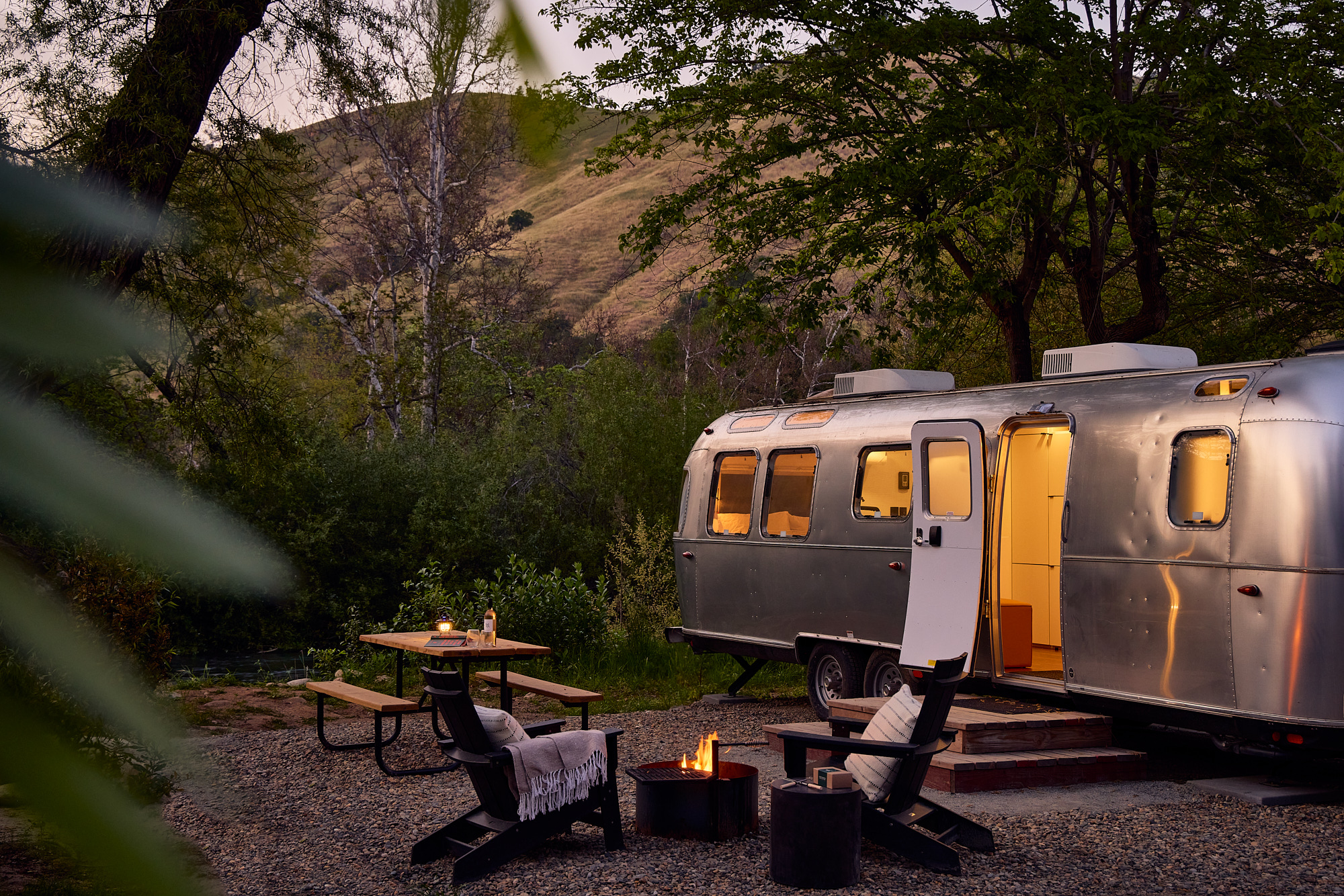 A polished silver Airstream trailer with a warmly lit interior sits in a peaceful canyon setting, surrounded by trees and hills, with outdoor lounge chairs, a picnic table, and a fire pit completing the glamping scene. Sage Outdoor Advisory includes high-end trailer experiences like this in Feasibility Studies to assess mobility, design appeal, and land use efficiency.