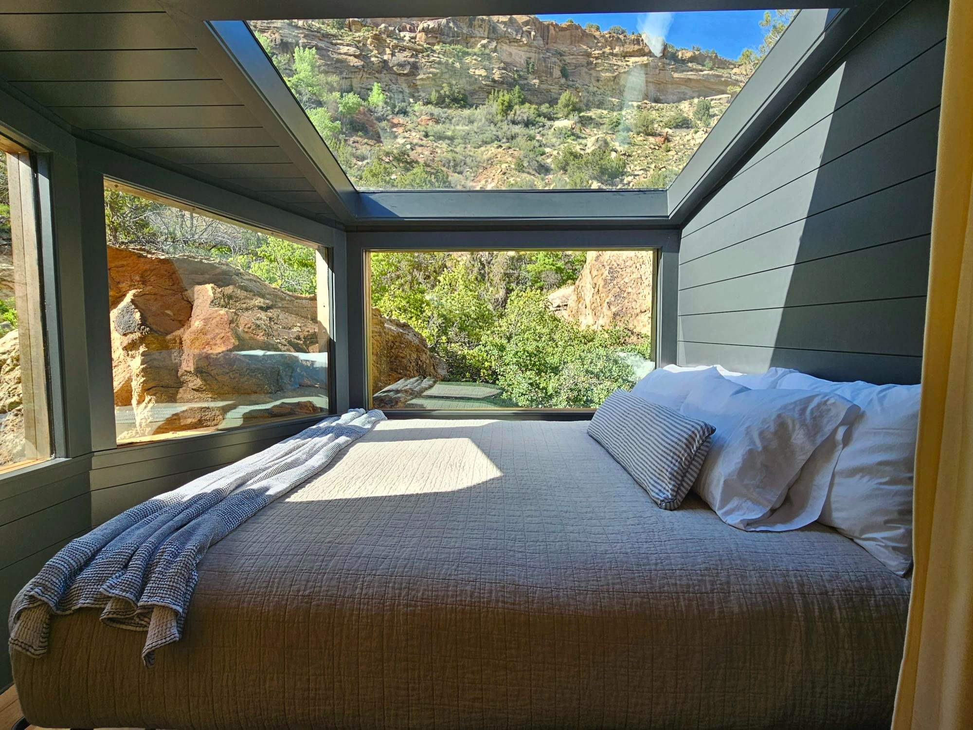 A cozy king-sized bed with a beige quilt and white pillows inside a modern glamping cabin, featuring floor-to-ceiling windows and a large skylight offering panoramic views of sandstone cliffs and desert vegetation. This immersive design, showcased in a Sage Outdoor Advisory Feasibility Study, highlights the luxury and nature-connected experiences central to glamping developments.