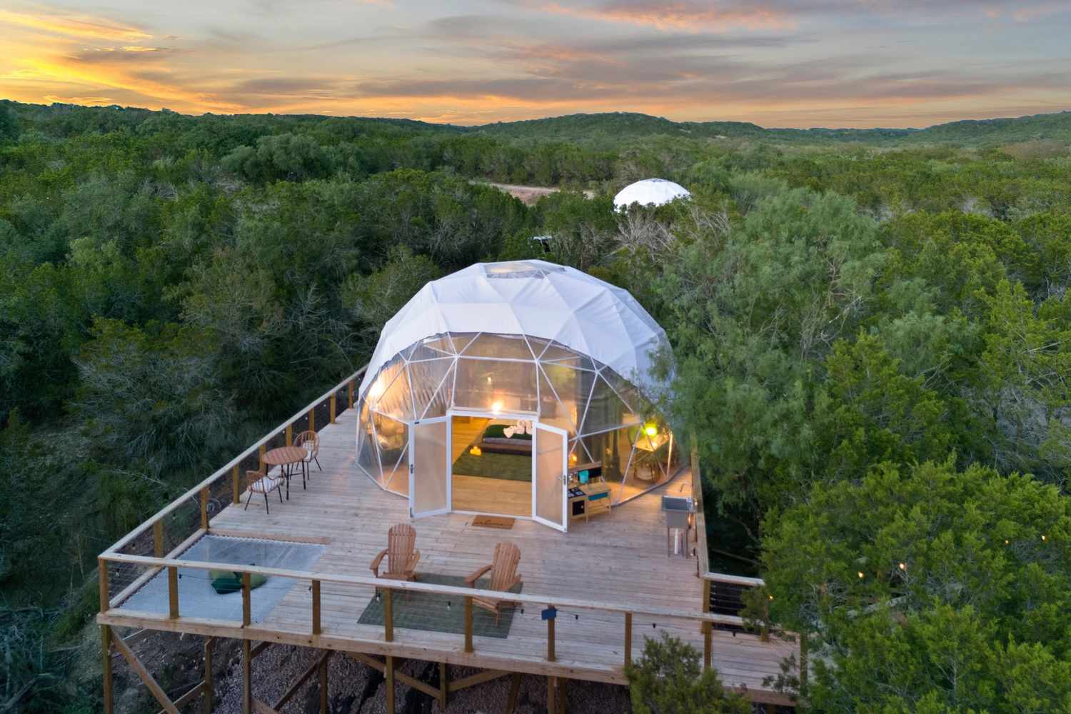 Beautifully designed transparent geodesic dome glamping structure perched atop an expansive wooden deck overlooking a serene, wooded landscape at sunset. Sage Outdoor Advisory's Feasibility Studies expertly evaluate the potential and viability of distinctive glamping properties such as this, ensuring successful ventures in outdoor hospitality.
