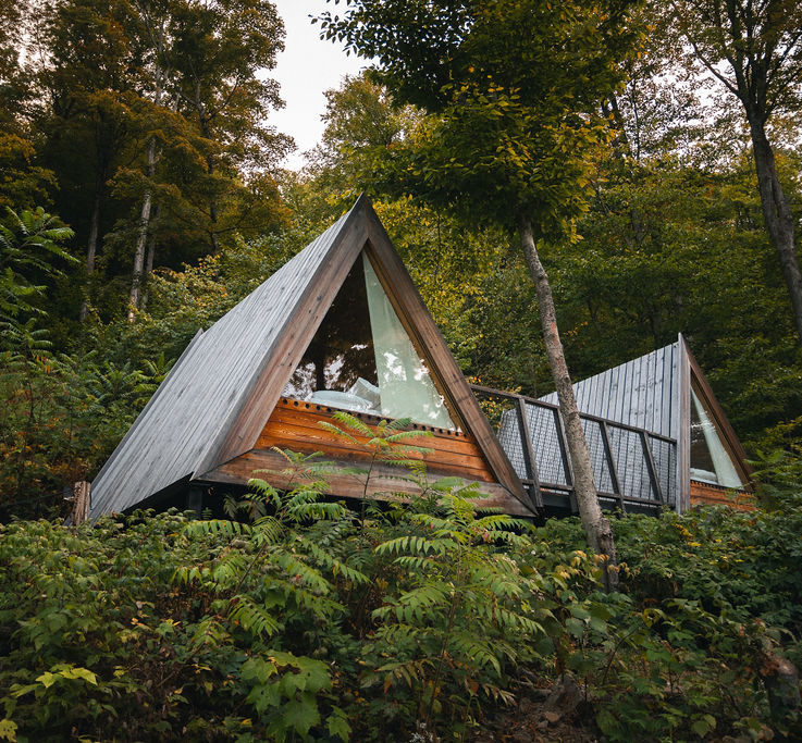 A pair of modern A-frame cabins with large triangular glass windows are nestled into a lush green hillside surrounded by dense forest, creating an immersive glamping experience. Sage Outdoor Advisory includes architecturally distinct units like these in Feasibility Studies to evaluate aesthetic appeal and terrain suitability in wooded environments.