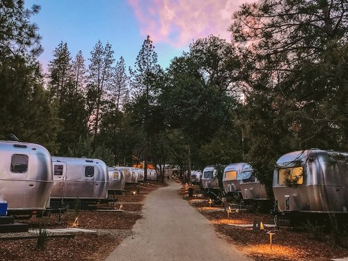 A line of gleaming Airstream trailers stretches along a forested path under a pastel dusk sky, creating a modern campground atmosphere nestled among tall pines. Sage Outdoor Advisory evaluates scalable, branded trailer-based glamping models like Autocamp in Feasibility Studies to guide regional expansion and guest capacity planning.
