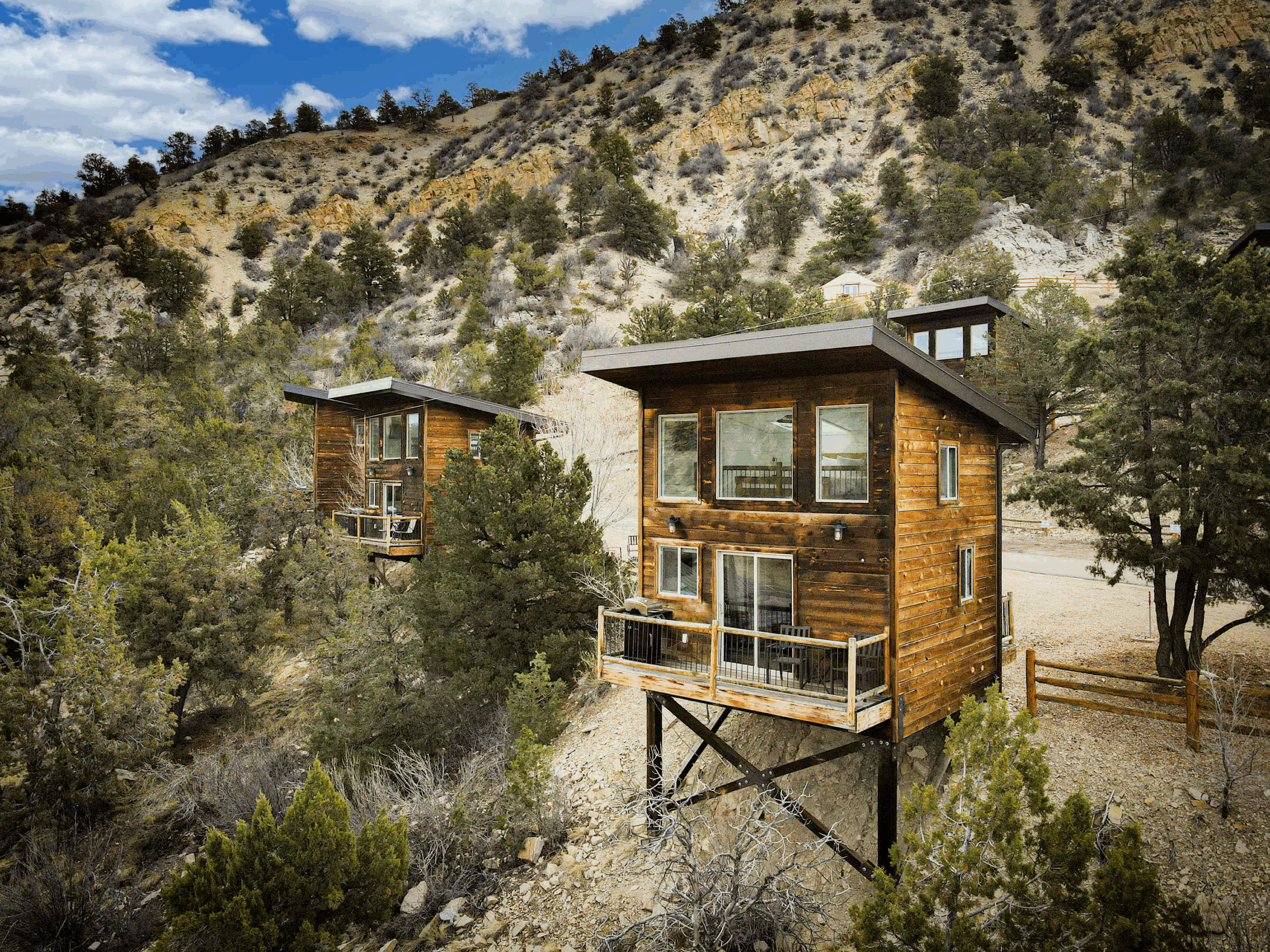 Modern two-story wood cabins perched on stilts along a rugged desert hillside, surrounded by juniper trees and dramatic canyon scenery. Sage Outdoor Advisory includes elevated glamping units like these in Feasibility Studies to assess terrain adaptability and panoramic view potential in mountainous regions.