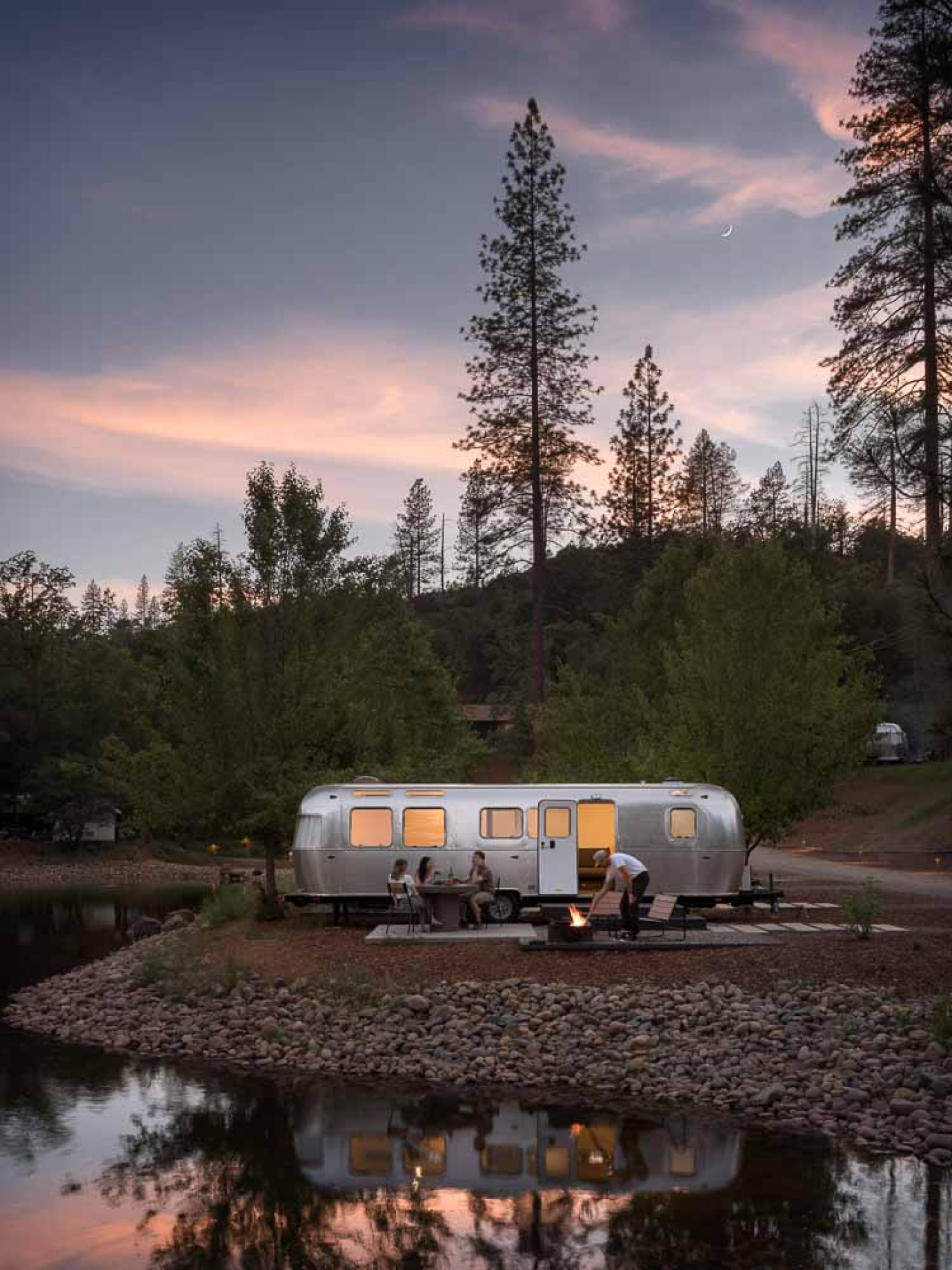 Scenic glamping setup with a modern Airstream trailer beside a tranquil pond at sunset, featuring guests enjoying a fire pit—an example of immersive outdoor hospitality evaluated in Sage Outdoor Advisory’s feasibility study for glamping resorts.