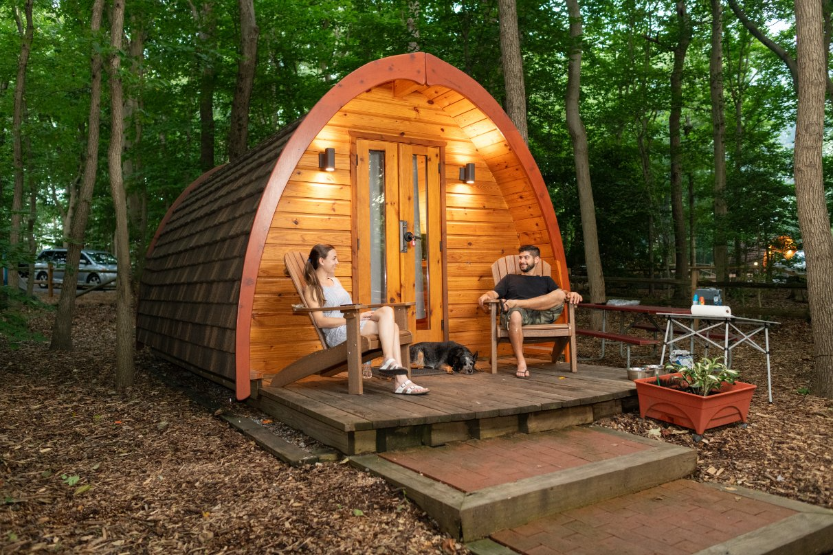 Two guests enjoying a relaxing evening on the front porch of a cozy wooden glamping pod nestled in a wooded setting, accompanied by their dog. Sage Outdoor Advisory regularly performs Feasibility Studies for charming, compact glamping structures to ensure successful guest experiences and investment returns.

