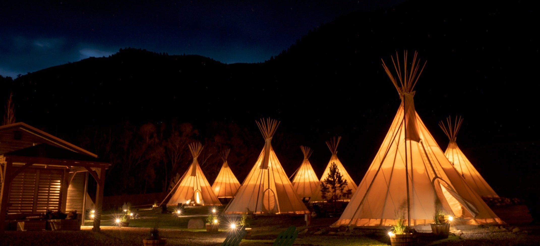 A cluster of softly illuminated tipis glows under a starry night sky, set against the backdrop of forested hills and deep wilderness at Dreamcatcher Tipi Hotel in Montana. Sage Outdoor Advisory includes culturally inspired glamping concepts like these in Feasibility Studies to assess experience-driven demand and site integration for boutique outdoor stays.