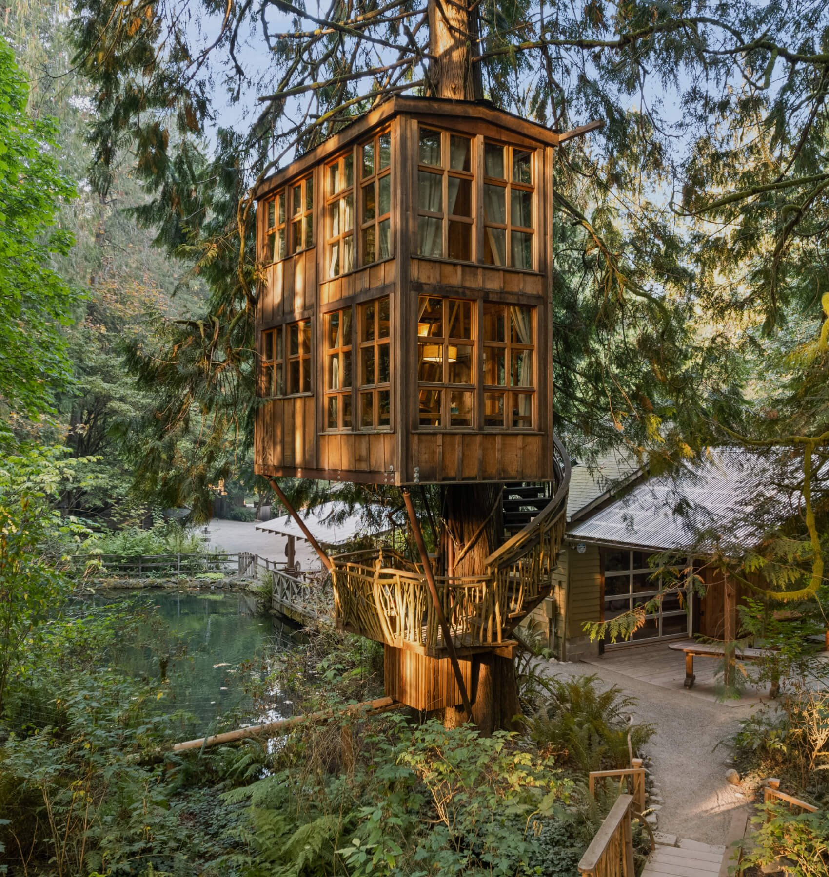 A multi-story treehouse built around a massive tree trunk features floor-to-ceiling windows and intricate wooden railings, perched above a lush forest floor and serene pond. Sage Outdoor Advisory analyzes elaborate, nature-integrated treehouses like this in Feasibility Studies to assess premium guest draw and land-use potential in eco-tourism projects.