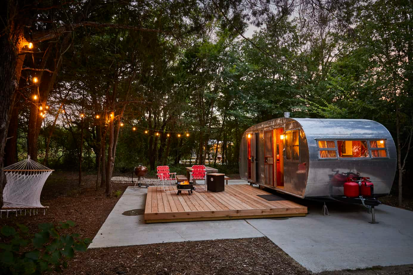 A polished silver vintage trailer sits on a raised wooden platform surrounded by twinkle lights, red lounge chairs, a hammock, and a cozy fire pit nestled among trees. Sage Outdoor Advisory includes retro trailer concepts like this in Feasibility Studies to assess nostalgic design appeal and return on investment in glamping properties.

