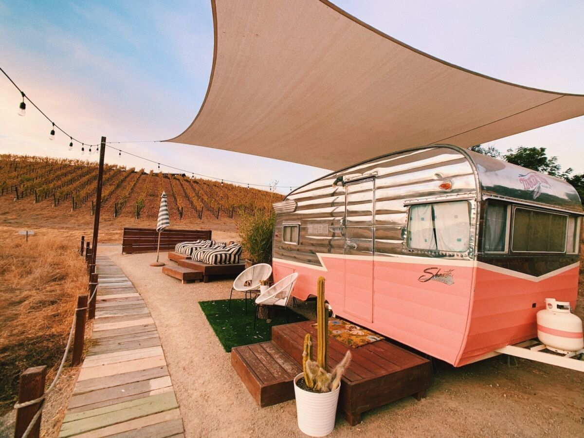 A shiny pink-and-silver vintage trailer sits beside a vineyard under a stretched canvas shade, with a boardwalk, cactus planter, outdoor chairs, and striped loungers completing the cozy glamping setup. Sage Outdoor Advisory includes retro-inspired trailers in Feasibility Studies to assess visual branding, guest experience, and integration with agritourism destinations.
