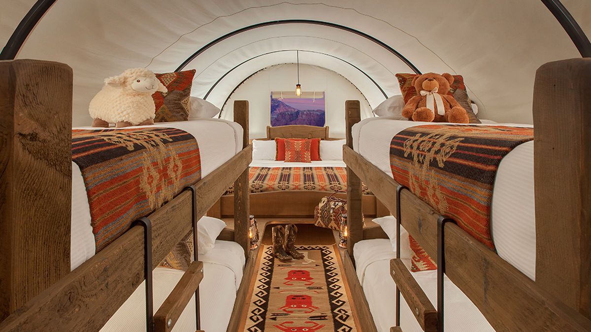 Interior of a luxury covered wagon glamping unit featuring a king bed and two sets of bunk beds with rustic wood frames, adorned with Southwestern textiles and plush toys. Sage Outdoor Advisory includes multi-bed layouts like this in Feasibility Studies to evaluate family accommodations and group retreat potential.