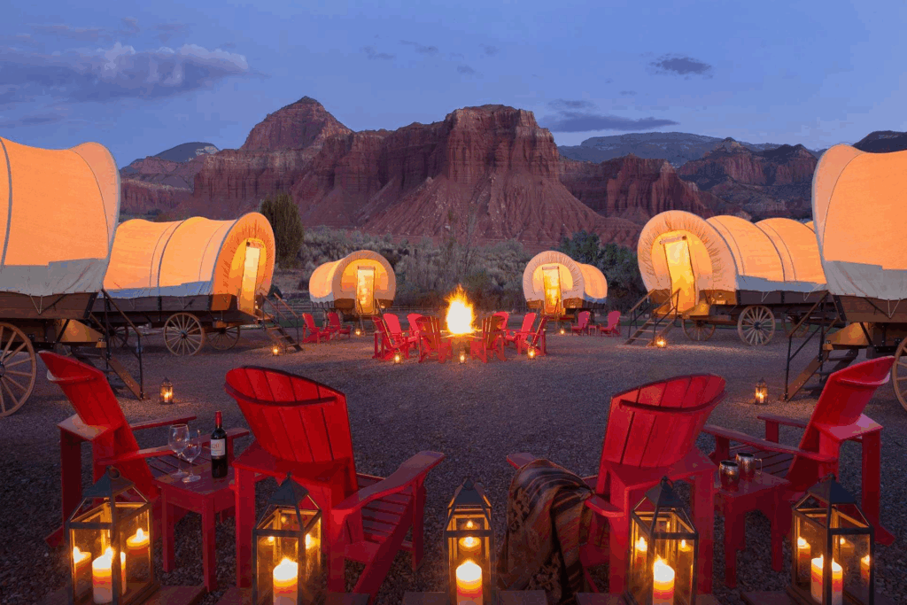 A circle of illuminated covered wagons forms a communal glamping space around a glowing campfire, set against dramatic red rock formations at dusk. Sage Outdoor Advisory analyzes culturally themed units like these in Feasibility Studies to guide experience-driven glamping development in iconic U.S. landscapes.