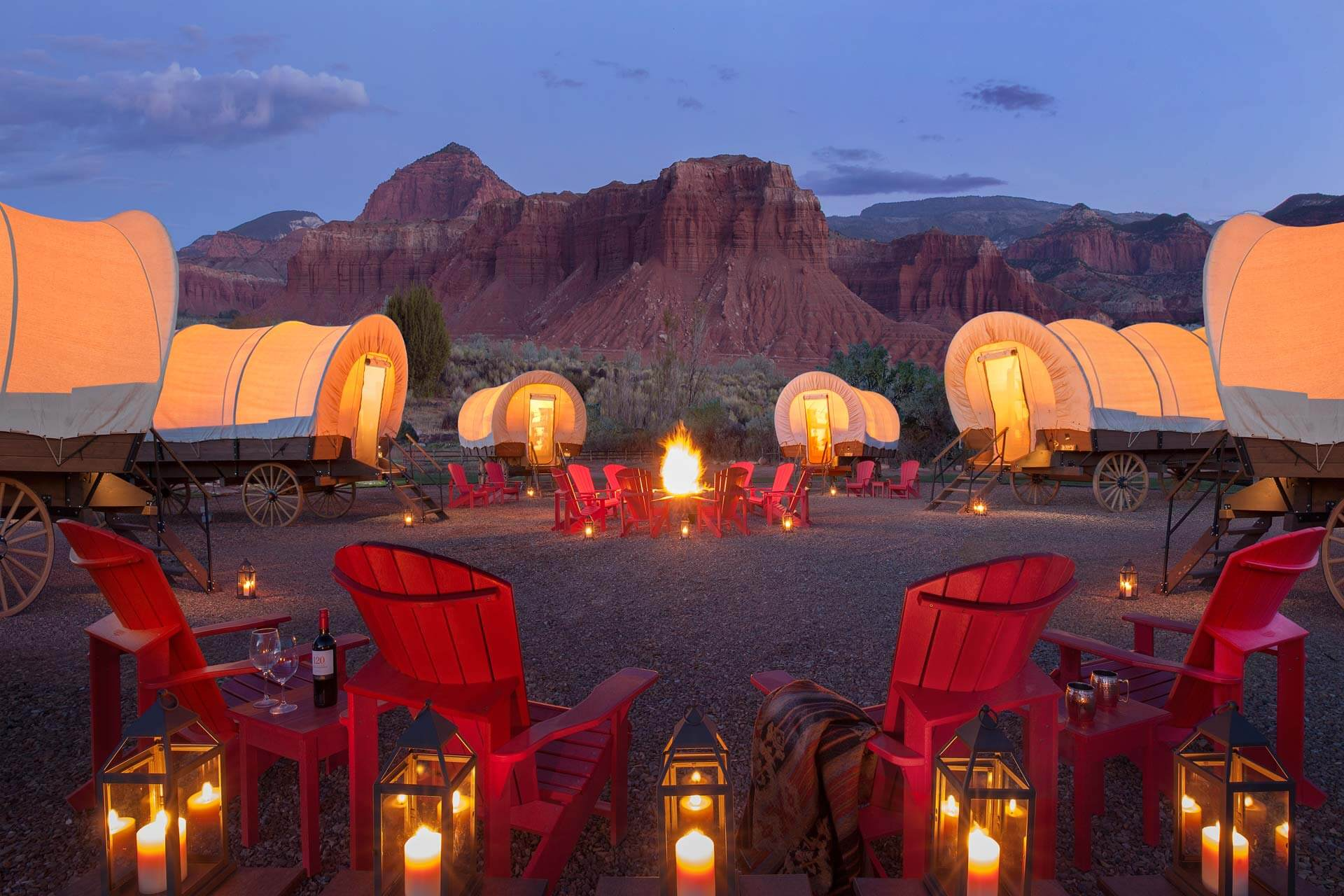 A circle of illuminated covered wagons forms a communal glamping space around a glowing campfire, set against dramatic red rock formations at dusk. Sage Outdoor Advisory analyzes culturally themed units like these in Feasibility Studies to guide experience-driven glamping development in iconic U.S. landscapes.