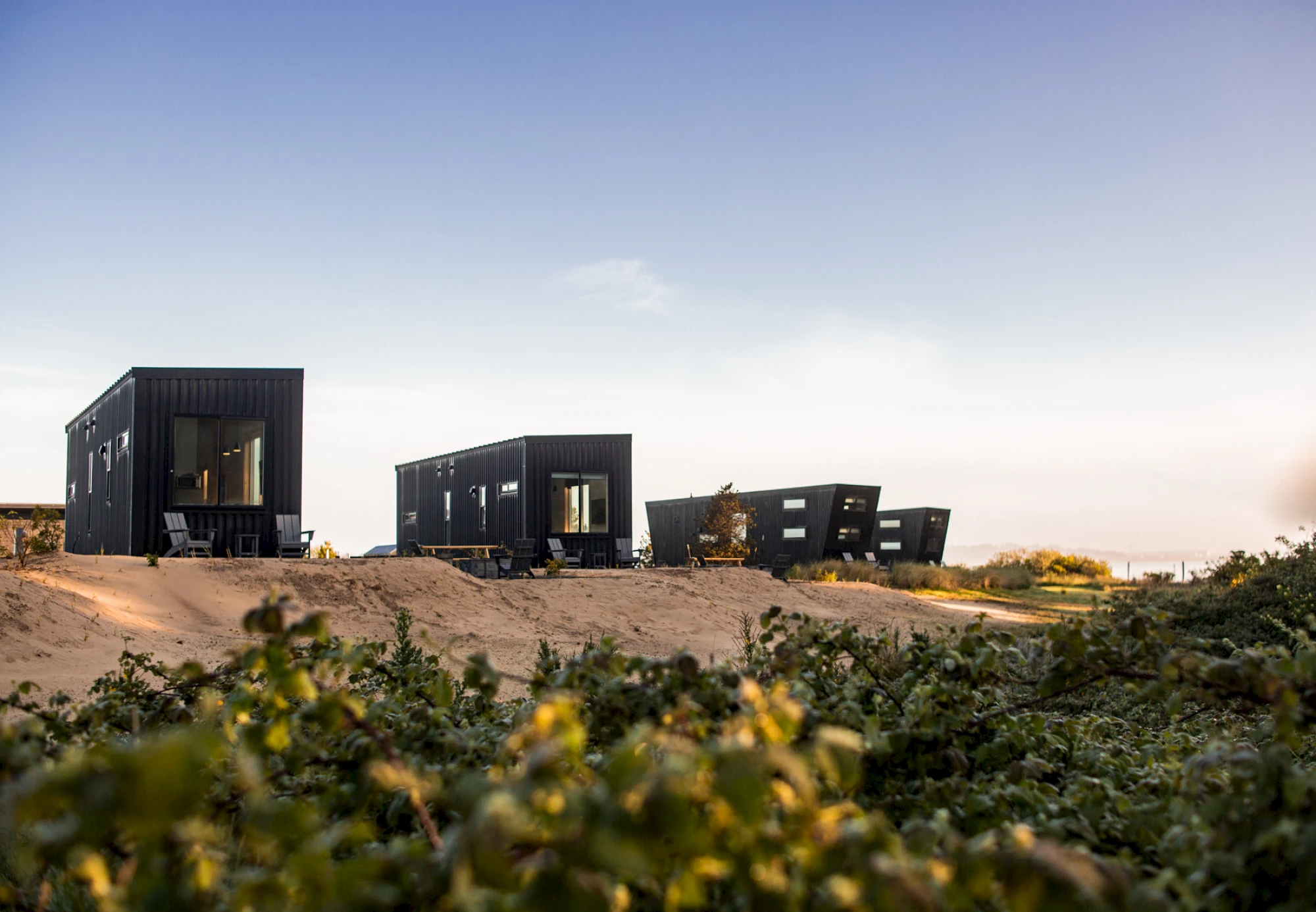 Modern black cabins on sandy dunes with outdoor seating, blending minimalist architecture with a natural coastal landscape. Sage Outdoor Advisory provides feasibility studies, appraisals, and consulting to ensure glamping and outdoor resort projects like this are both visionary and financially viable.