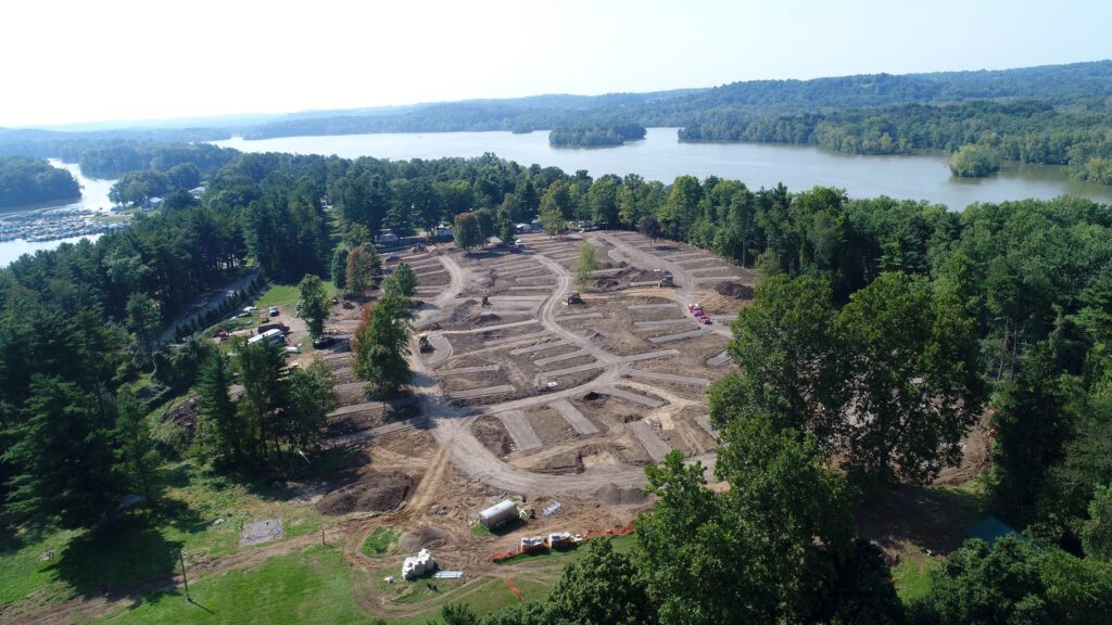 Aerial view of Charles Mill Lake Park Beach Campground in Mansfield, Ohio during construction, showing newly graded RV site pads, curved internal roadways, and earthwork in progress, surrounded by dense green forest with the lake visible in the background.