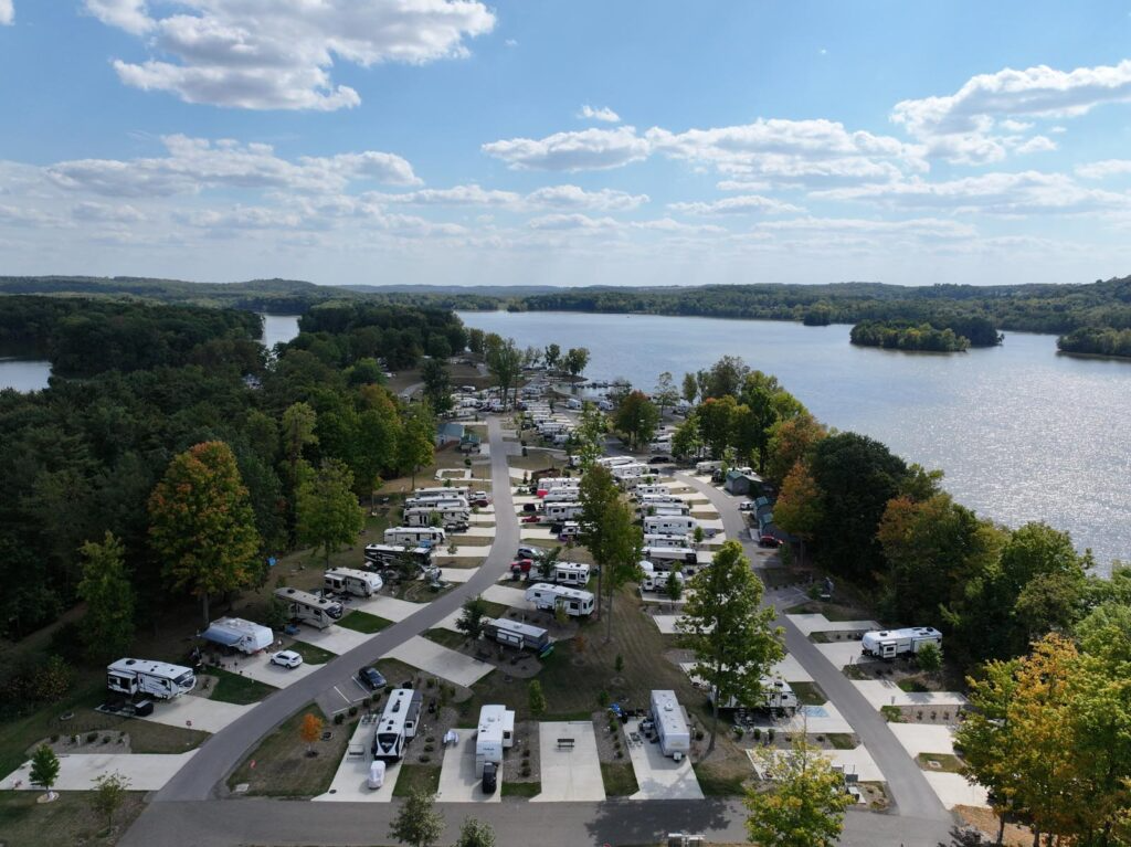 Aerial view of the completed Charles Mill Lake Park RV campground with paved roads, organized concrete pads, parked RVs, and landscaped site areas, set among wooded shoreline terrain with expansive lake views under a bright blue sky.