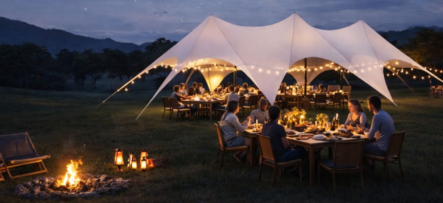 Outdoor evening dining event under a large white stretch tent with multiple peaks, illuminated by string lights. Two long wooden tables with chairs are set up underneath, with people gathered for dinner. A small fire pit and lanterns glow in the foreground, with mountain silhouettes and a starry twilight sky in the background.
