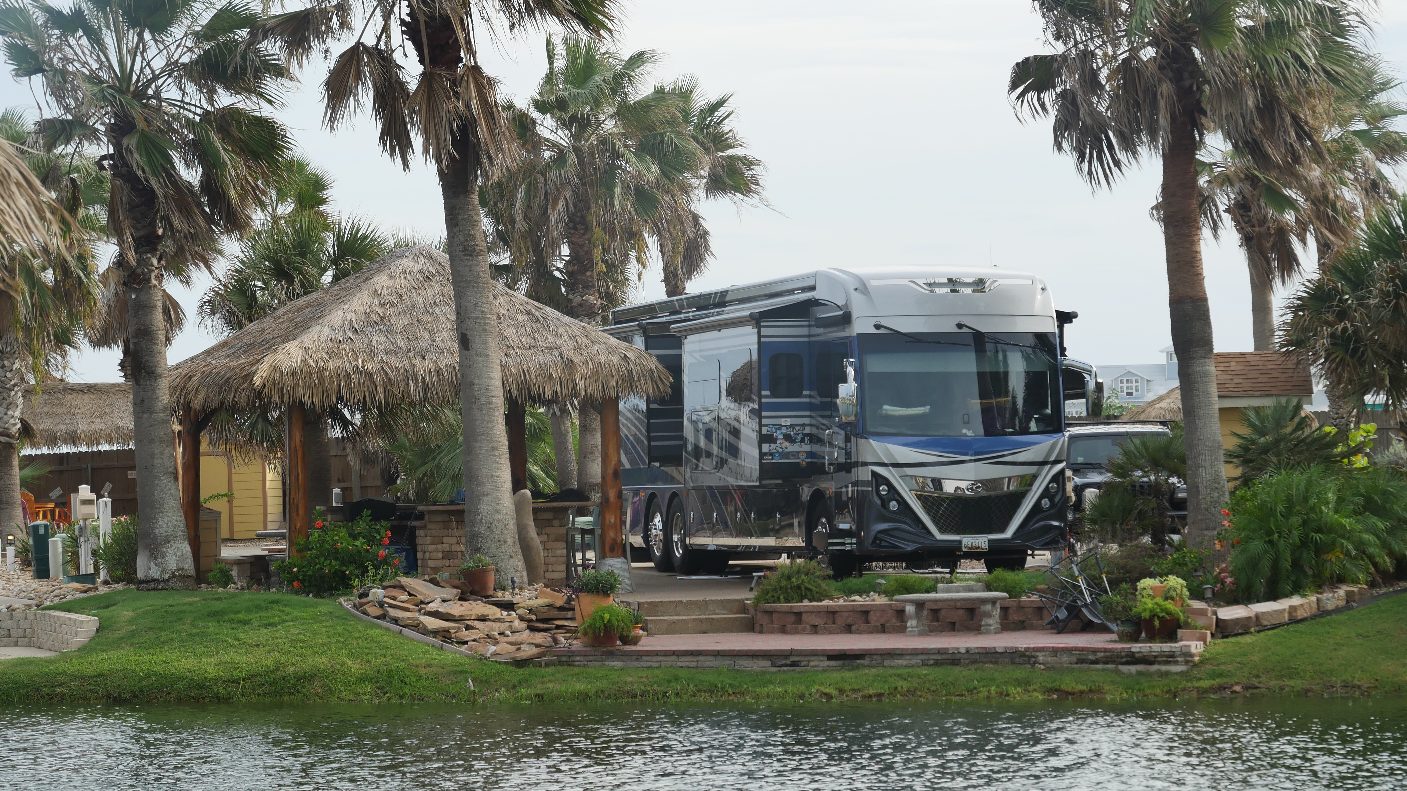 Waterfront RV site at a full service RV resort, featured in the Sage Outdoor Advisory Partner Spotlight. A large Class A motorhome in blue, silver, and gray sits on a concrete pad with its slide out and awning extended, framed by tall palm trees under an overcast sky. To the left, a thatched palapa on rough timber posts shelters an outdoor kitchen counter and bar stools. The site is finished like a small yard: a paver patio, stacked stone retaining walls, a stone bench, landscaped beds with potted plants and flowering shrubs, and steps down to a strip of mown grass along the water in the foreground. Yellow outbuildings and a light blue two story house are visible in the background.